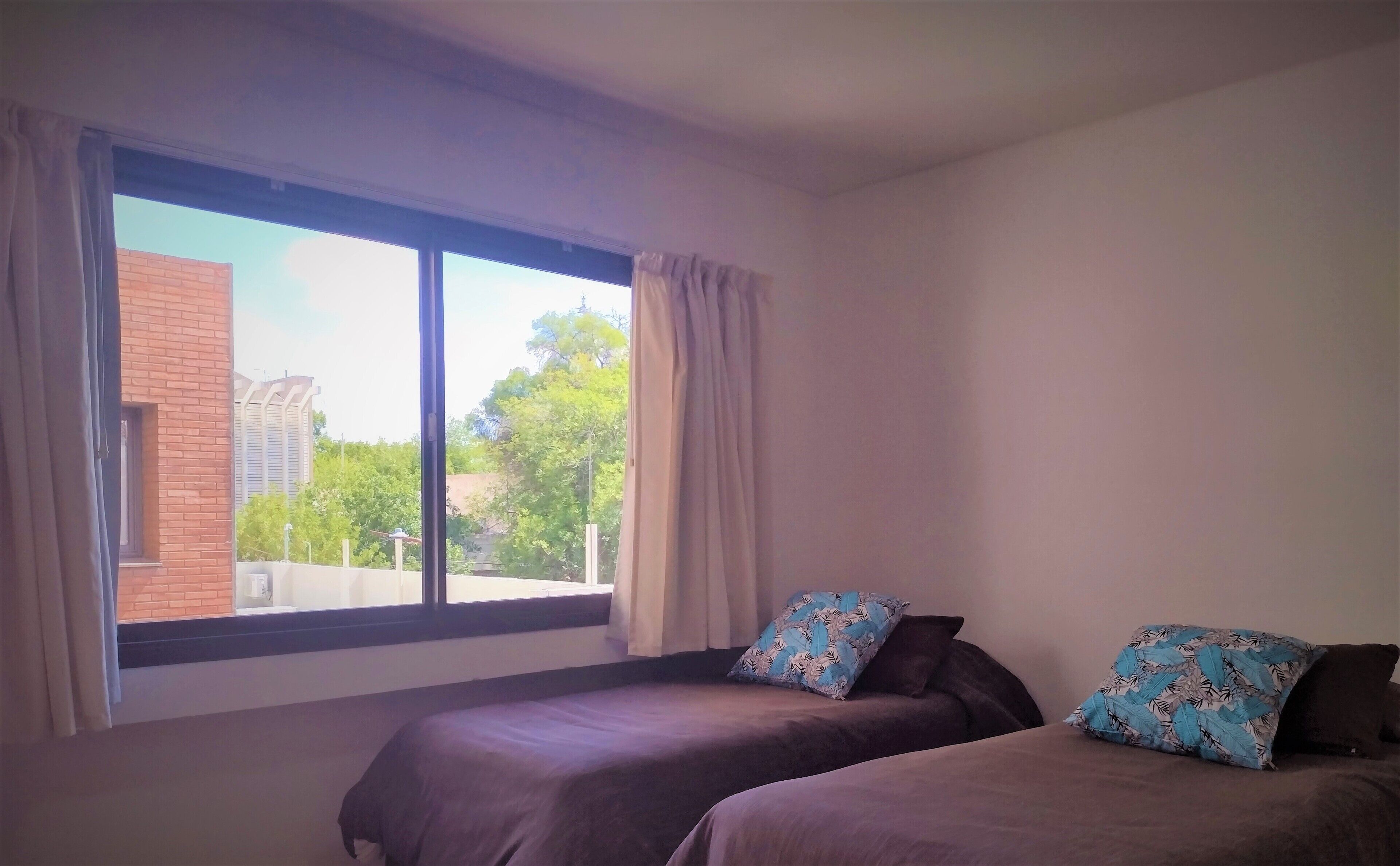 A room with two beds, a large window with curtains, and a view of buildings and trees outside.