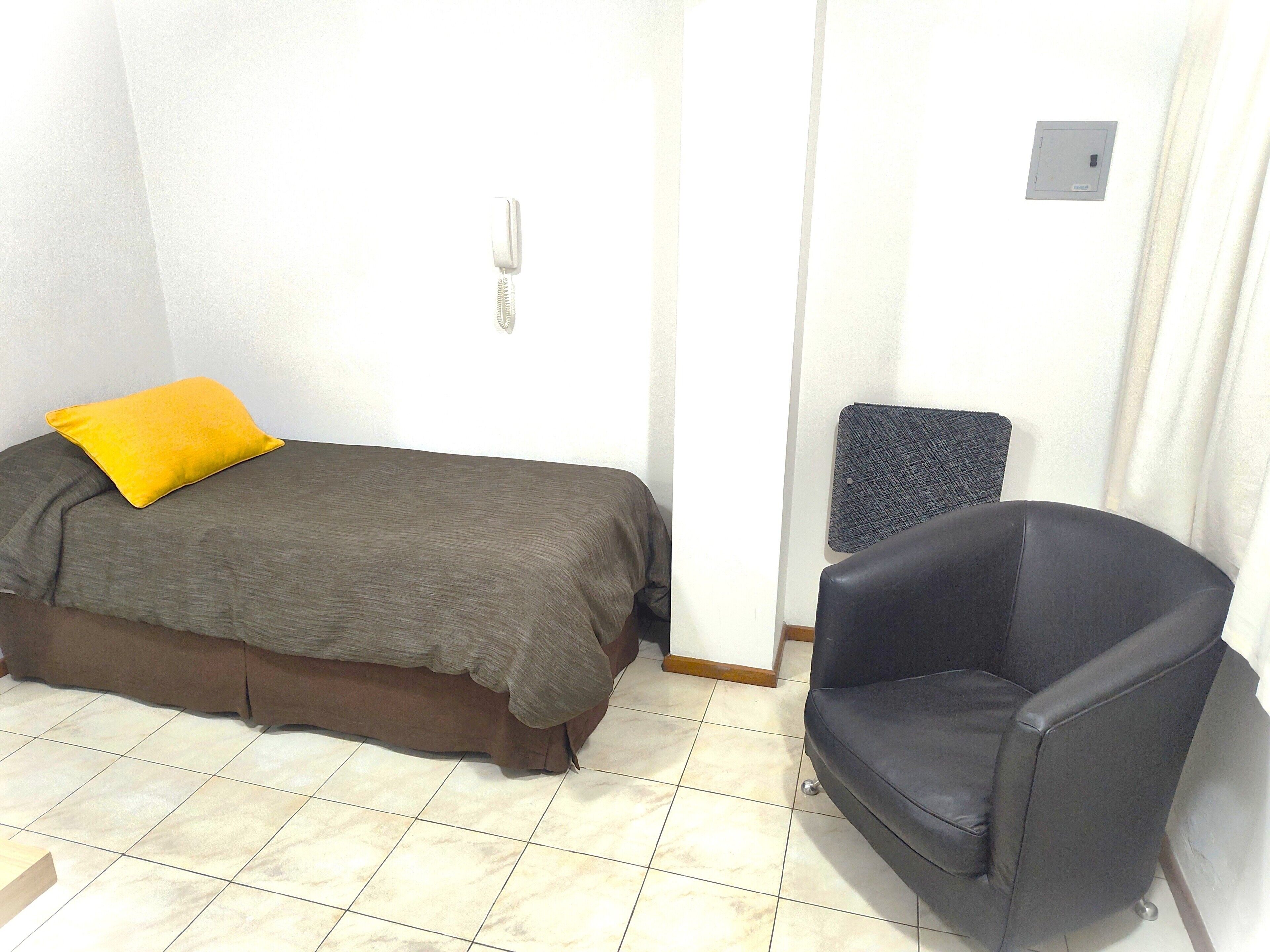 A small, sparsely furnished room with a bed, a chair, and a wall-mounted light switch.