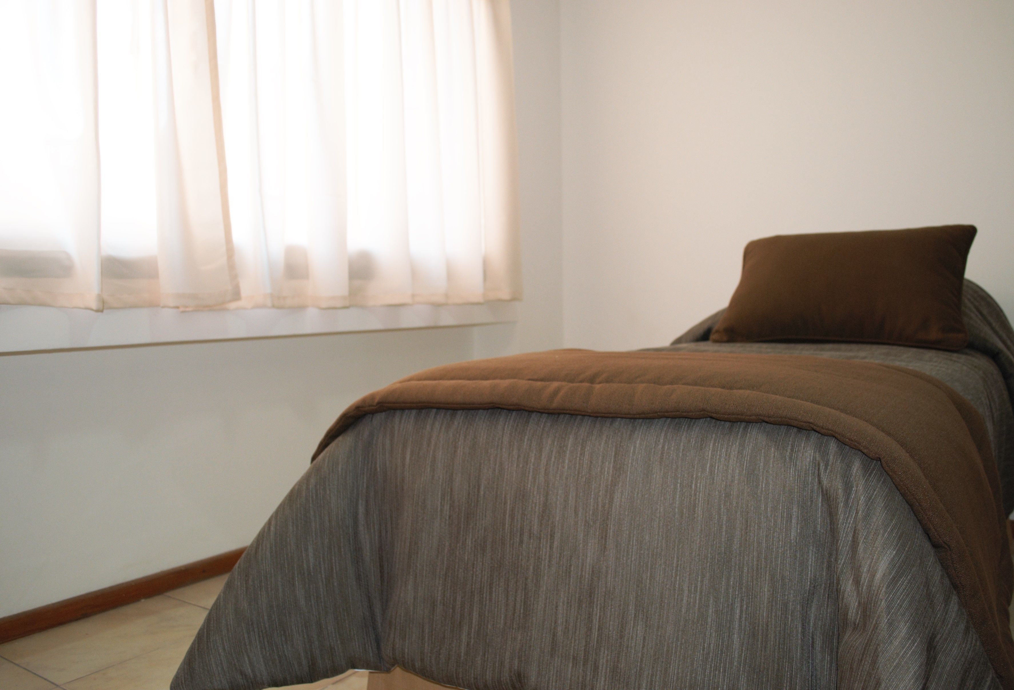 A bedroom with a bed, a window with sheer curtains, and a brown pillow.
