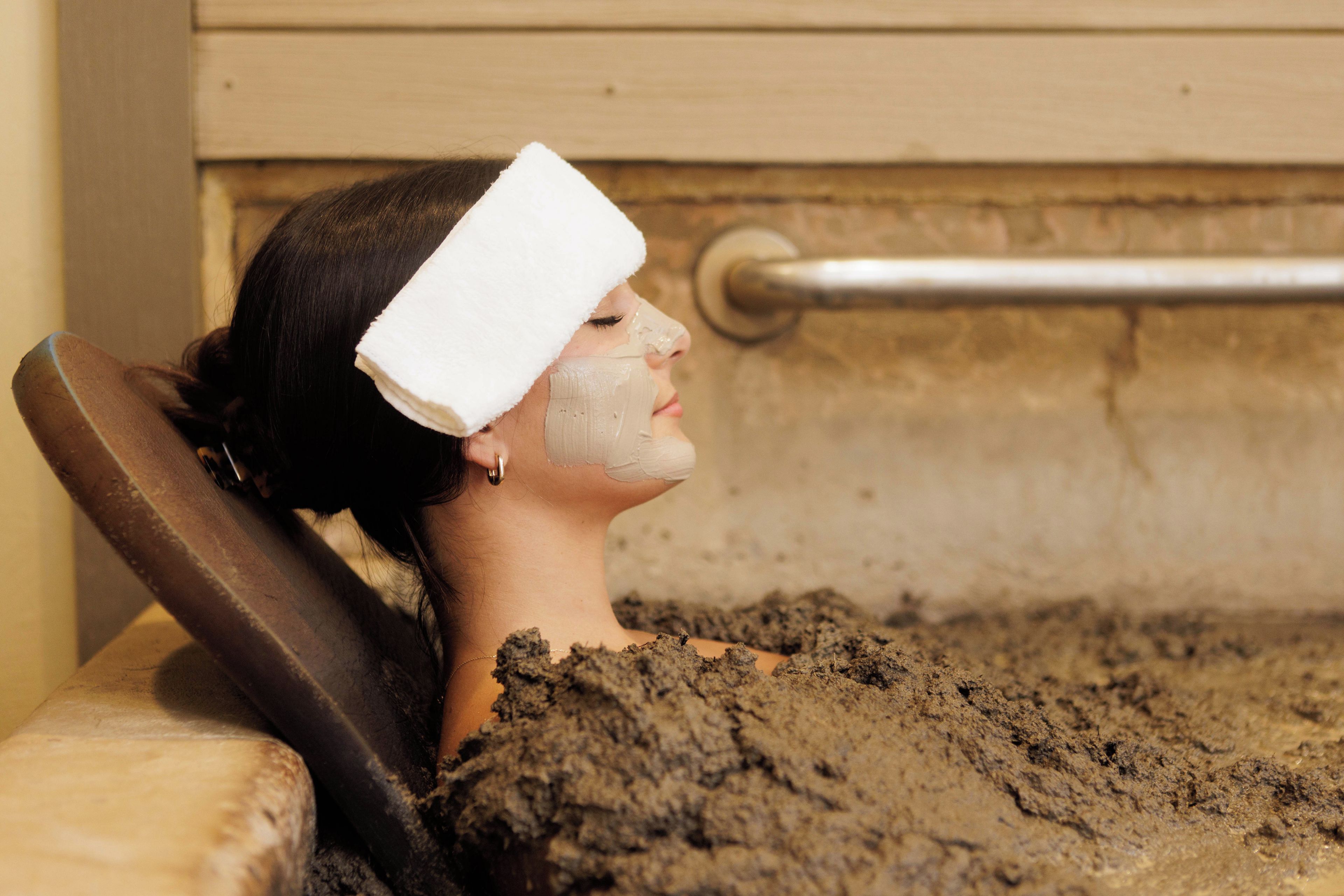 Une personne allongée dans une baignoire remplie de boue, avec une rambarde en métal et un mur en bois en arrière-plan.