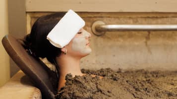 Une personne allongée dans une baignoire remplie de boue, avec une rambarde en métal et un mur en bois en arrière-plan.