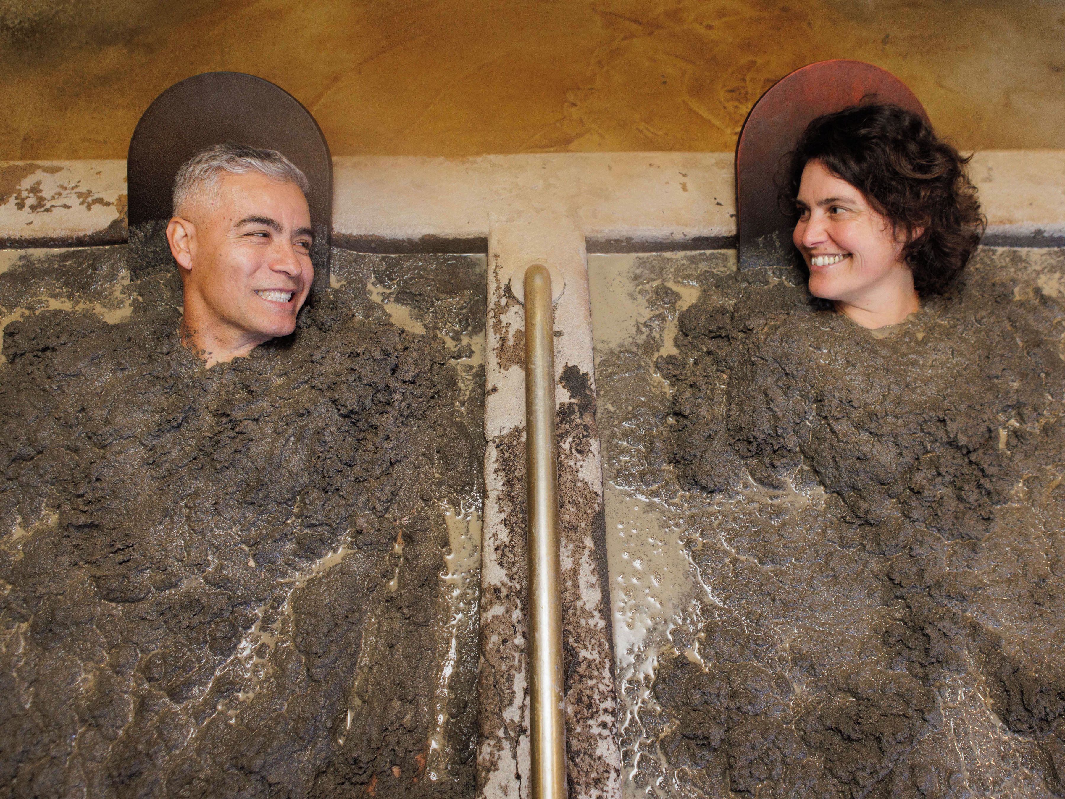 Two individuals in a bathtub filled with mud, with a metal bar in the center.