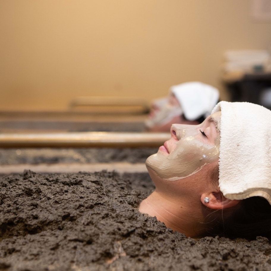 A person lying in a spa bath filled with mud, with another person in the background.