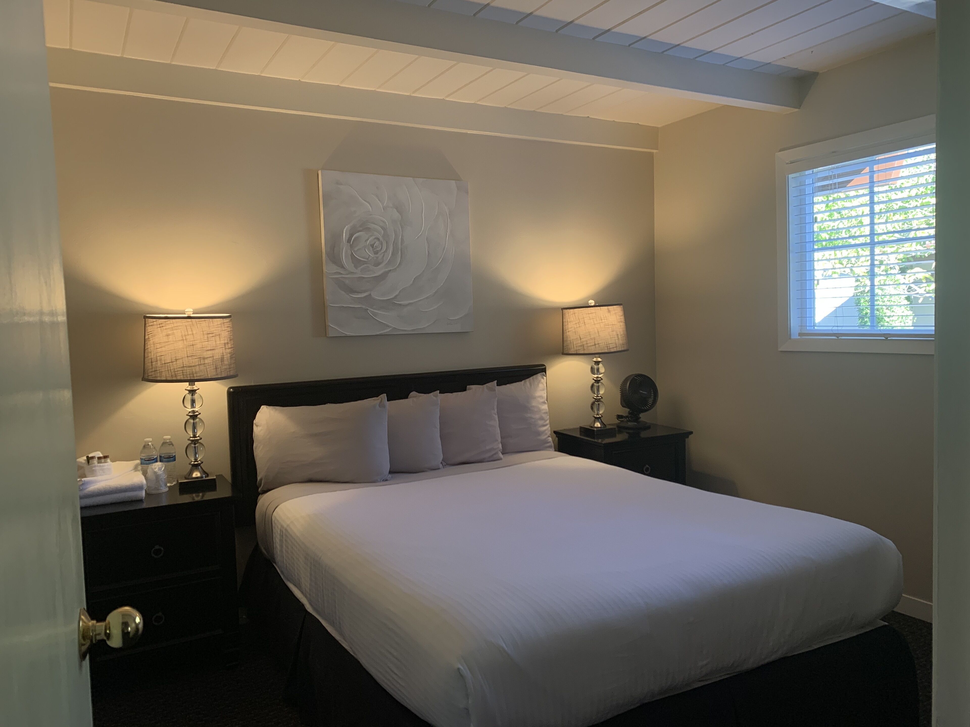 A neatly made bed with white linens, two bedside tables with lamps, a wall-mounted artwork, and a window with blinds.
