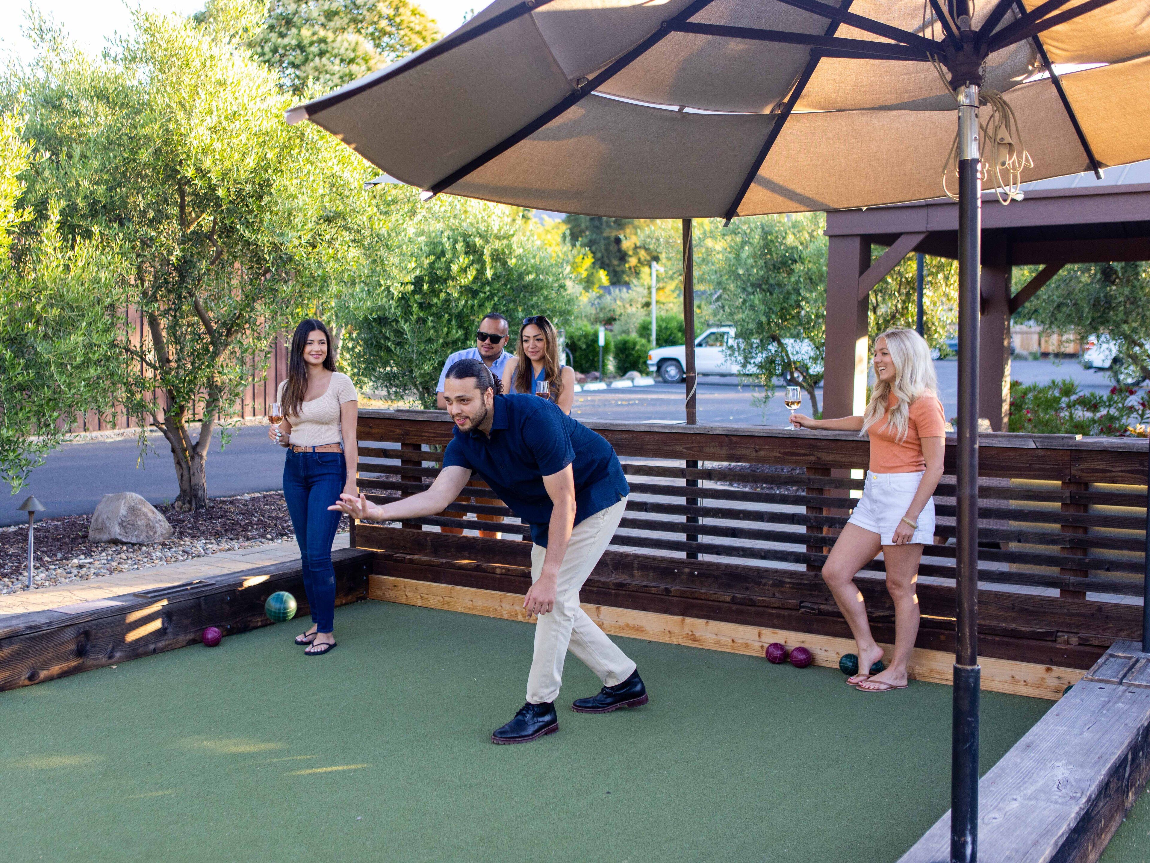 A group of people playing croquet on a green field with a wooden bench and an umbrella.