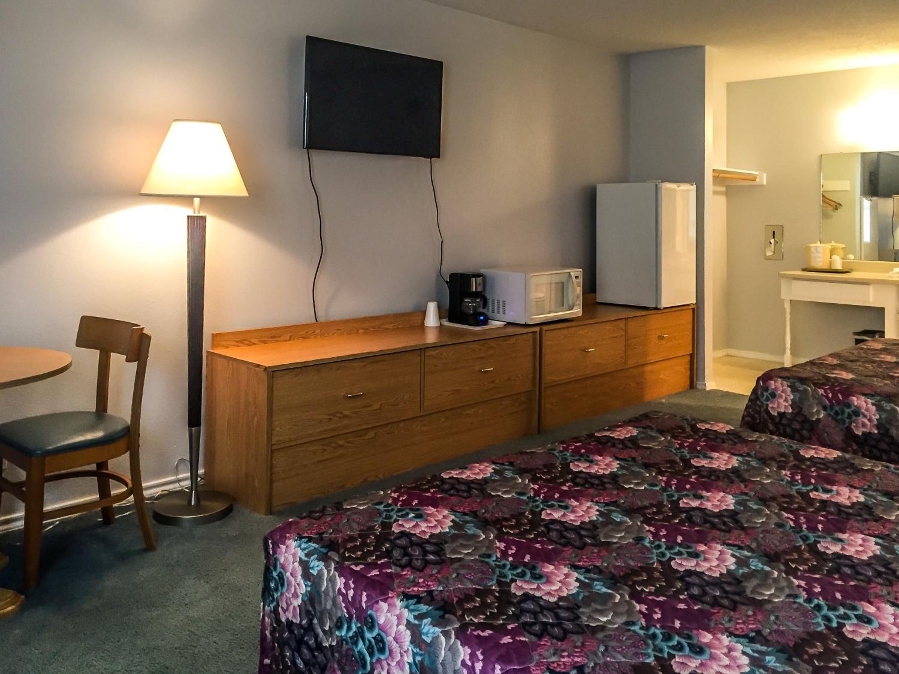 Une chambre d’hôtel équipée d’un bureau en bois, d’une chaise, d’un lit avec une literie à motifs floraux, d’une télévision, d’un four à micro-ondes et d’une petite kitchenette avec un réfrigérateur.