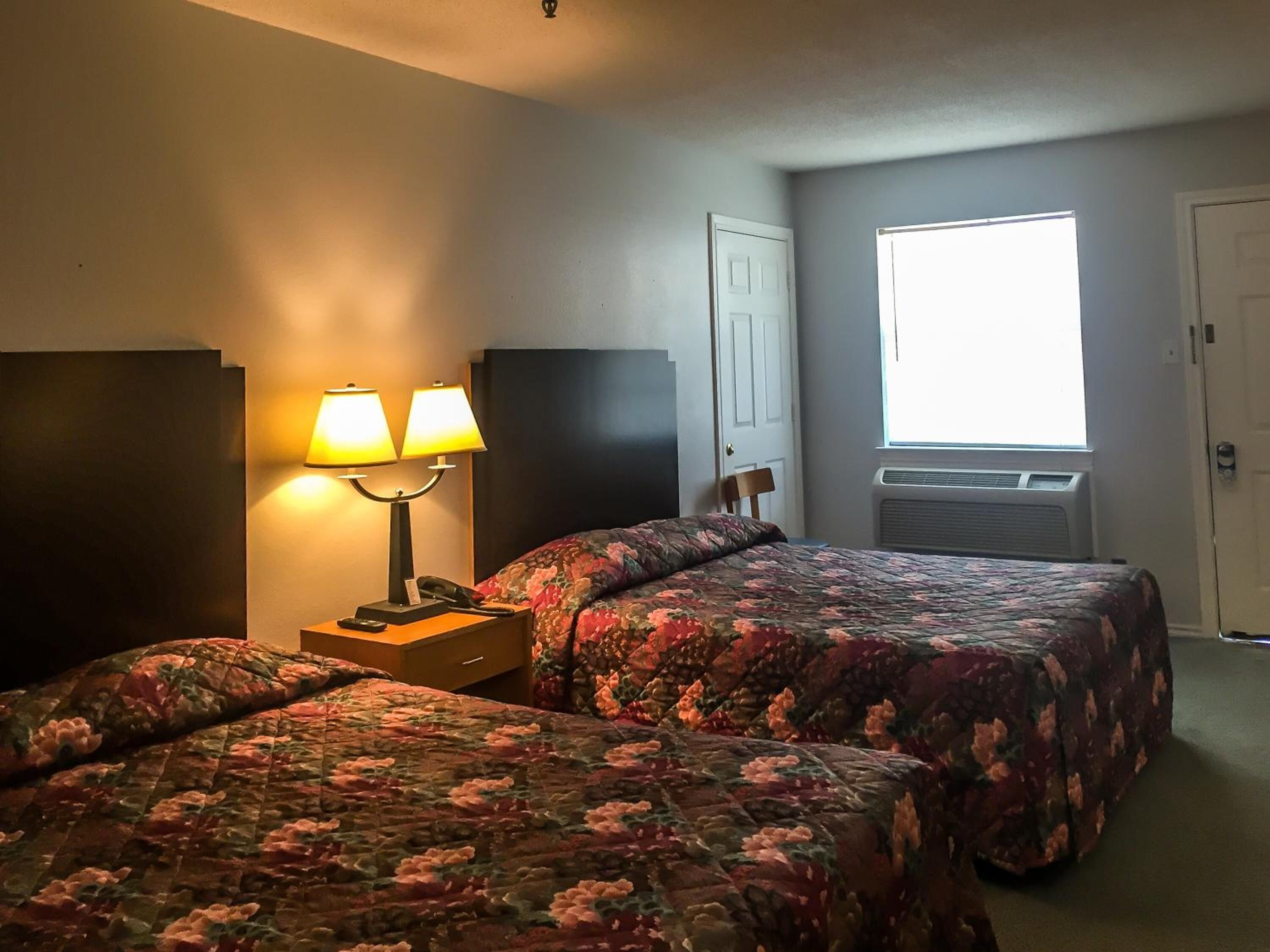 Una habitación de hotel con dos camas, una mesita de noche con lámpara, una ventana con persianas y un aire acondicionado.