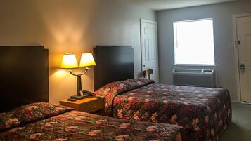 Una habitación de hotel con dos camas, una mesita de noche con lámpara, una ventana con persianas y un aire acondicionado.