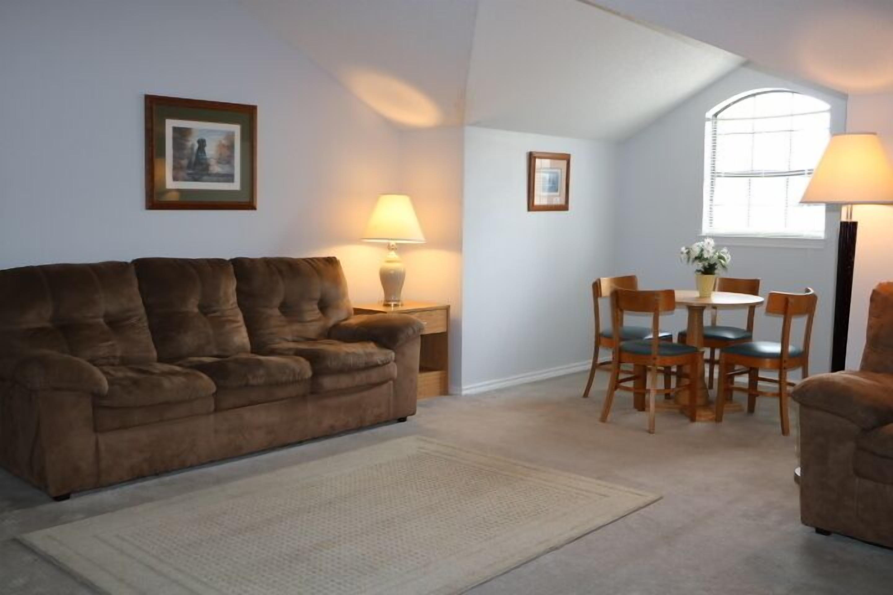 A living room with a brown sofa, a small table with chairs, a lamp, and framed pictures on the wall.
