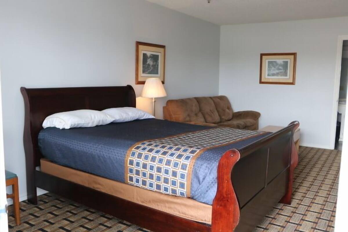 Una cama bien hecha con estructura de madera, una colcha azul y blanca estampada y una única almohada blanca. Hay una silla tapizada marrón con un cojín, una mesita con una lámpara y dos cuadros enmarcados en la pared. El suelo tiene un diseño de baldosas estampado.