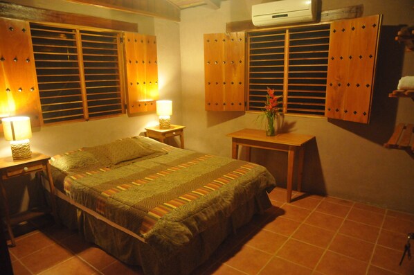 Un dormitorio con cama, mesitas de noche, una mesa y una ventana con persianas de madera.