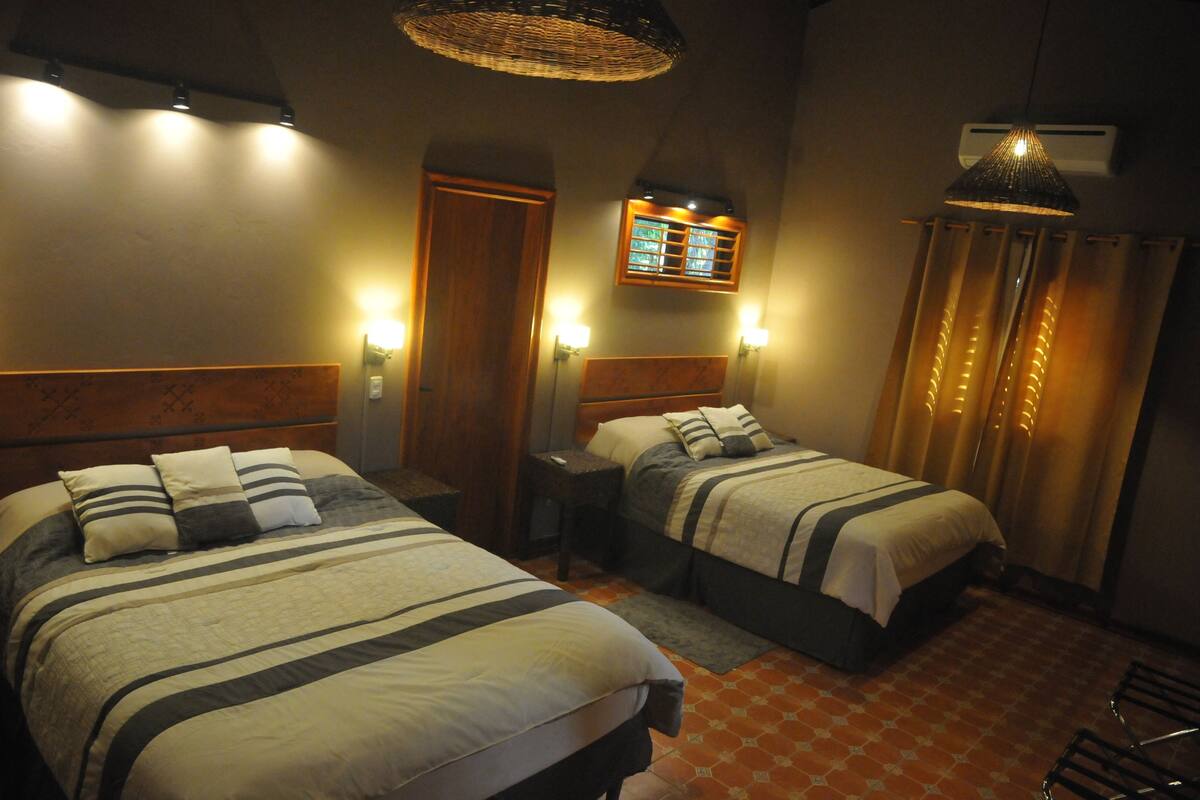 A hotel room with two beds, a small table, a chair, a wall-mounted air conditioner, and a window with curtains.