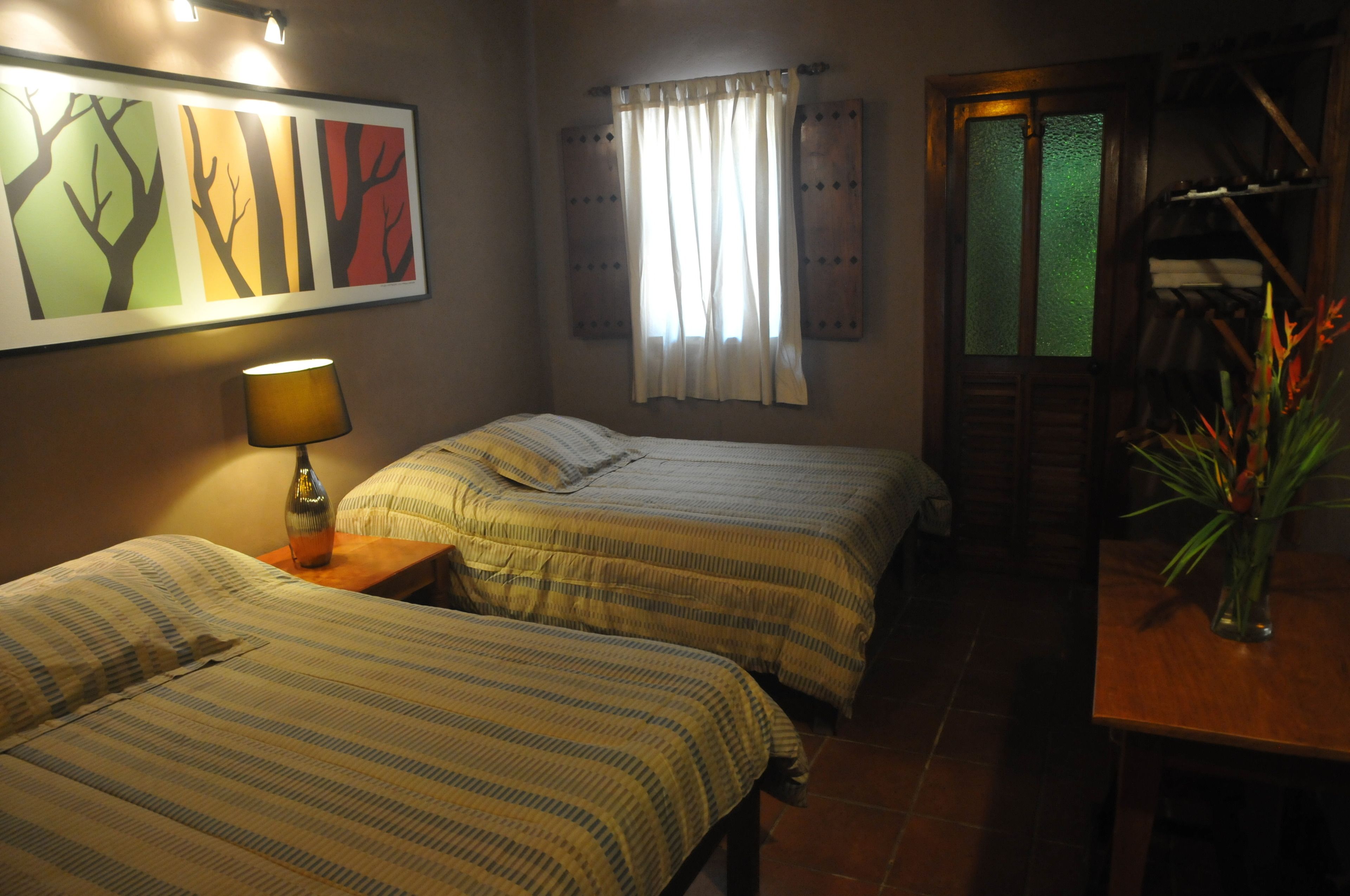 A room with two beds, a lamp, a painting, a window with curtains, and a wooden table with a vase.