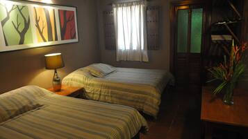 A room with two beds, a lamp, a painting, a window with curtains, and a wooden table with a vase.
