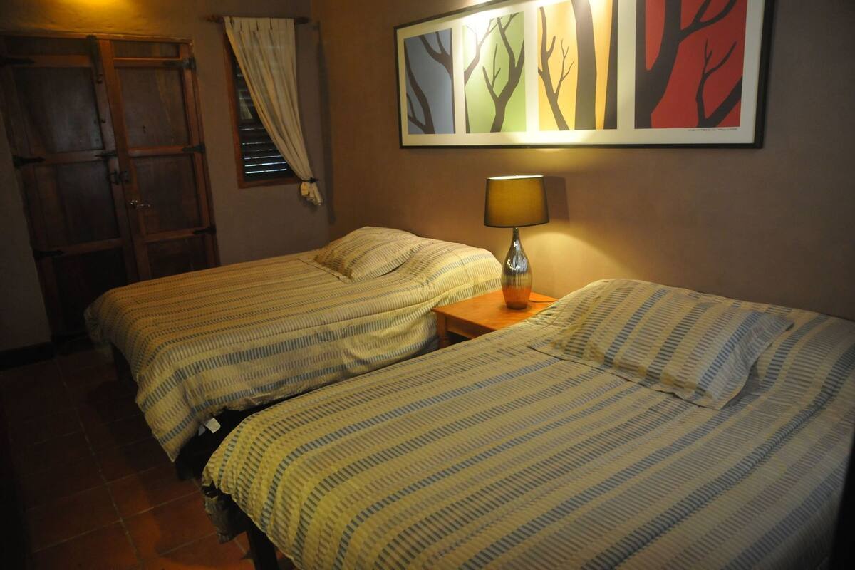 A room with two beds, a wooden door, a window with curtains, and a wall-mounted lamp.