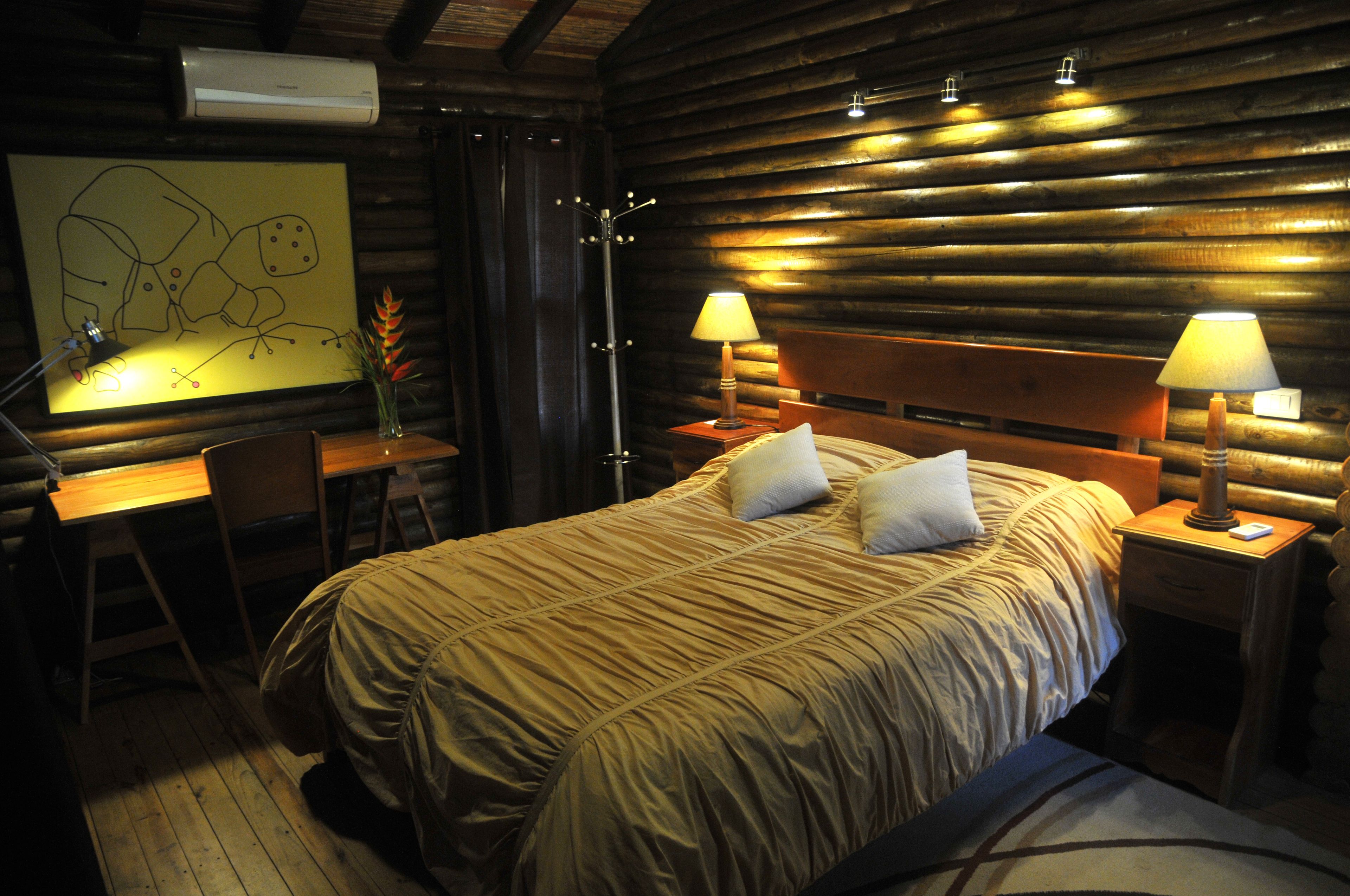 Acogedora habitación tipo cabaña de madera con una cama grande, dos lámparas de noche, un escritorio con silla y aire acondicionado instalado en la pared.