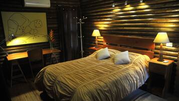 A cozy log cabin room with a large bed, two bedside lamps, a desk with a chair, and a wall-mounted air conditioner.