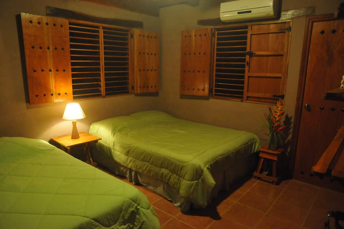 A room with two beds, wooden shutters, a small table with a lamp, and a potted plant.