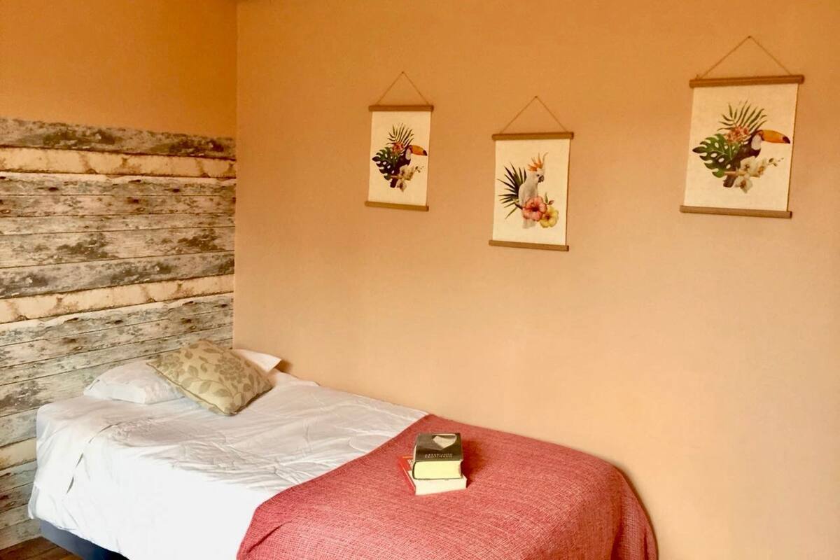 Une chambre à coucher avec une tête de lit en bois, un lit recouvert de draps blancs et trois œuvres d’art accrochées au mur, représentant des oiseaux et des fleurs tropicaux.