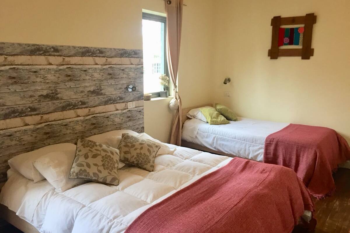 Une chambre avec deux lits, dont l’un est doté d’une tête de lit en bois et l’autre d’une tête de lit simple. On y trouve une fenêtre avec des rideaux, une horloge murale et un tableau encadré accroché au mur.