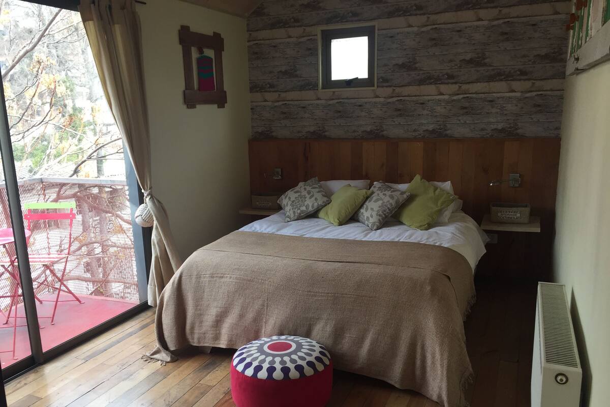 Une chambre à coucher avec une tête de lit en bois, un lit agrémenté de plusieurs oreillers, un pouf rouge et une petite fenêtre.