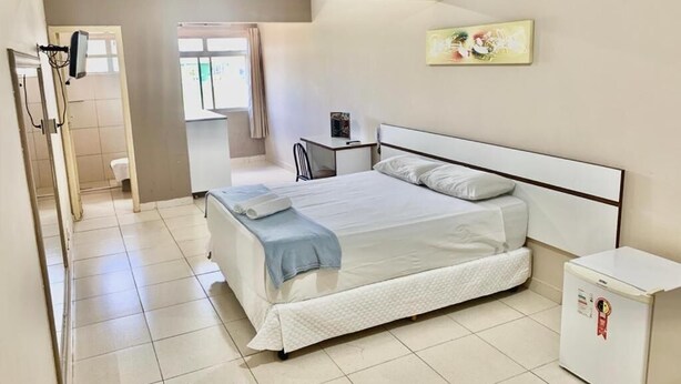 A bedroom with a bed, a bedside table, a small cabinet, and a window. - Dok Brasília Hotel - Unidade Taguatinga (Águas Claras)