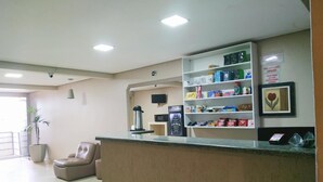 A reception area with a counter, a coffee machine, a refrigerator, a TV, and a shelf with various items. - Dok Brasília Hotel - Unidade Taguatinga (Águas Claras)