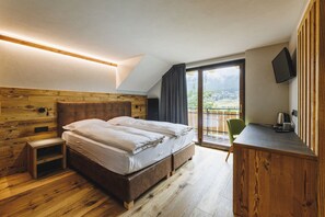A bedroom with a wooden headboard, a bed with white bedding, a small wooden nightstand, a desk with a chair, and a television on the wall. - Hotel Steinbock (Livigno)