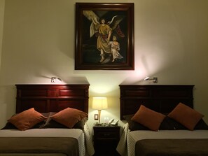 A hotel room with two beds, a nightstand with a lamp, and a painting of an angel above the beds. - Hotel San Xavier (Querétaro)