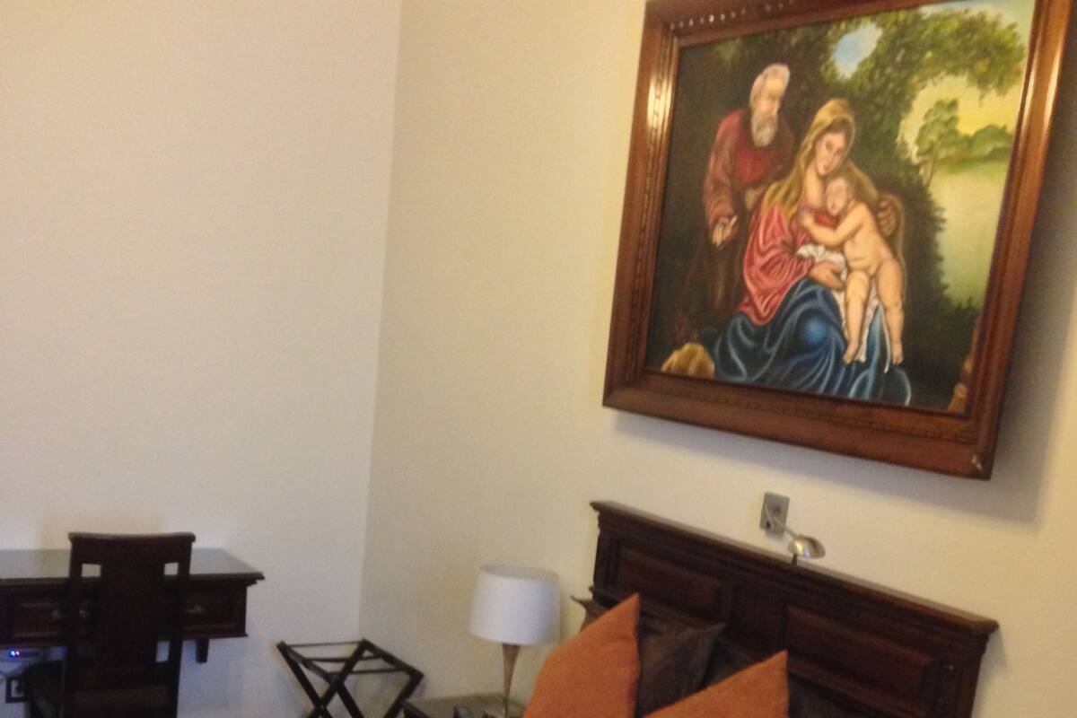 A hotel room with a bed, a desk, a chair, a lamp, and a painting on the wall.