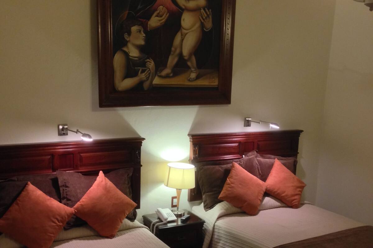 A hotel room with two beds, a nightstand with a lamp, a painting on the wall, and a wooden headboard.