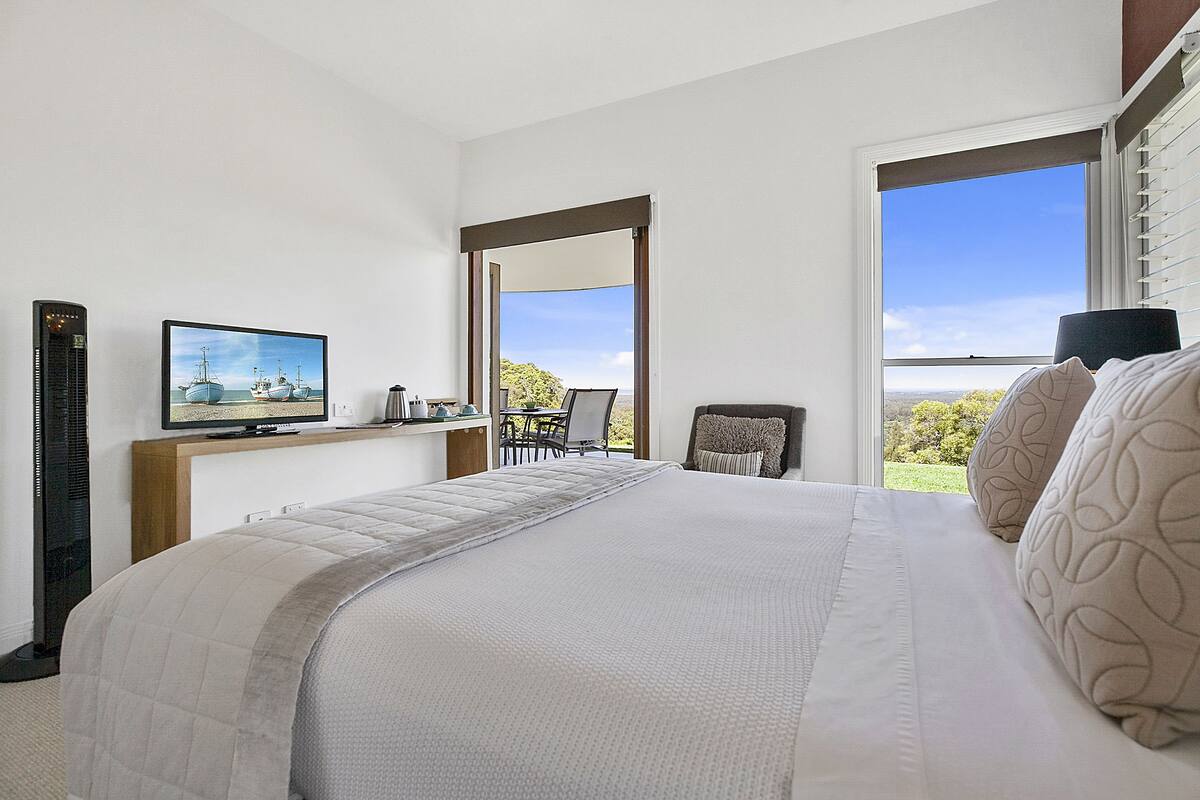A bedroom with a large bed, a television on a stand, a chair, and a window with a view of greenery.