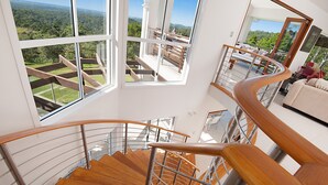 A modern interior with a spiral staircase, large windows offering a view of a lush landscape, and a balcony with outdoor furniture. - Alexandria Retreat (Doonan)