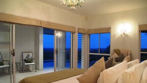 A bedroom with a large bed, a chair, and a view of the night sky through the windows. - Alexandria Retreat (Doonan)