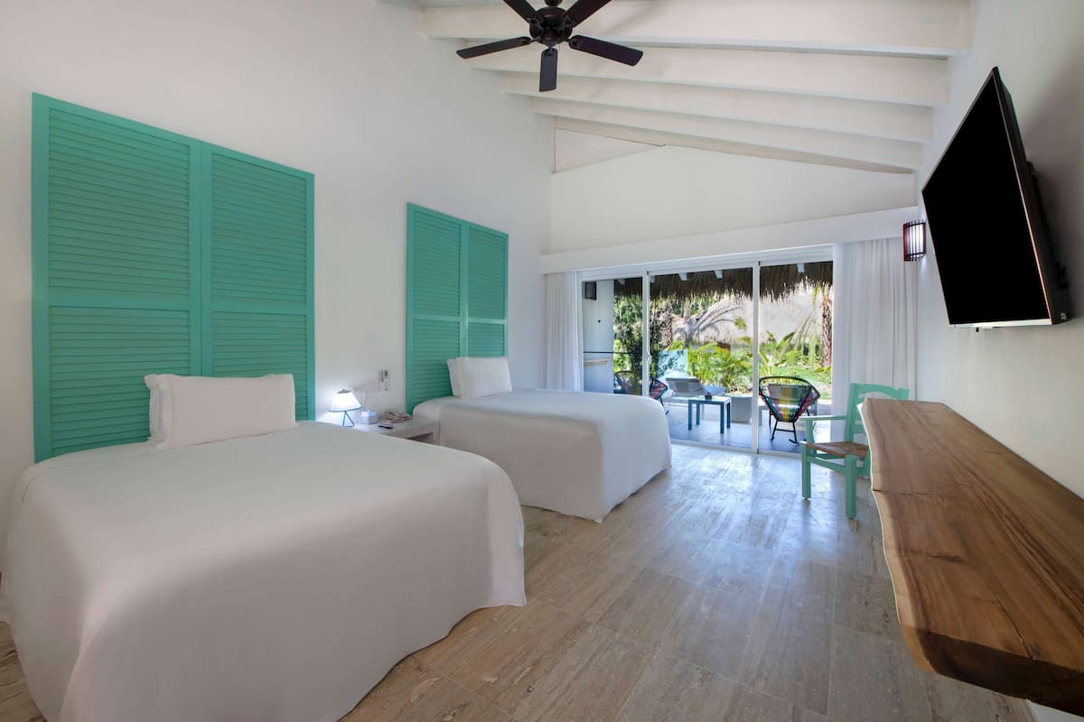 A hotel room with two beds, a ceiling fan, a TV, and a balcony with a view of greenery.
