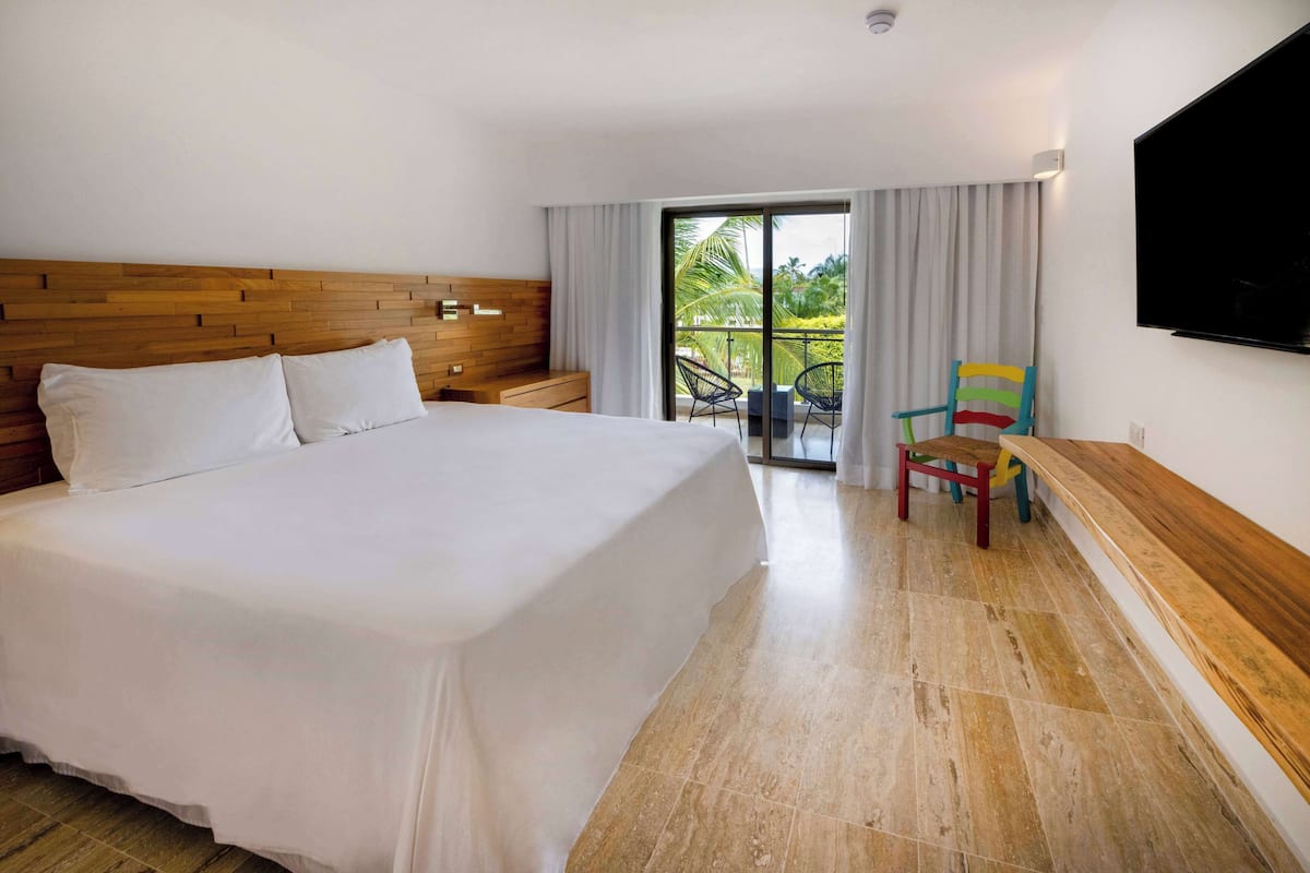 A hotel room with a large bed, a wooden headboard, a TV mounted on the wall, a balcony with a chair, and a wooden bench.