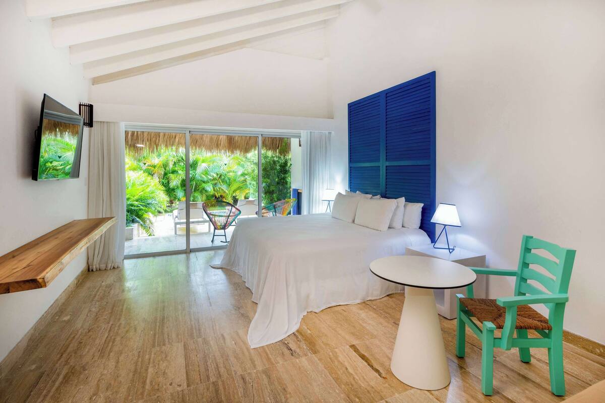 A bedroom with a large bed, a small table, a chair, a TV, and a balcony with a view of greenery.