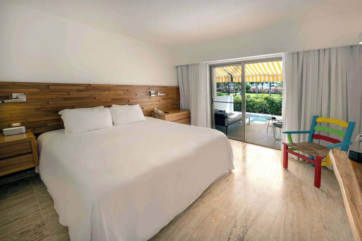 A hotel room with a large bed, wooden headboard, a nightstand, a chair, and a view of an outdoor area with a pool and greenery.