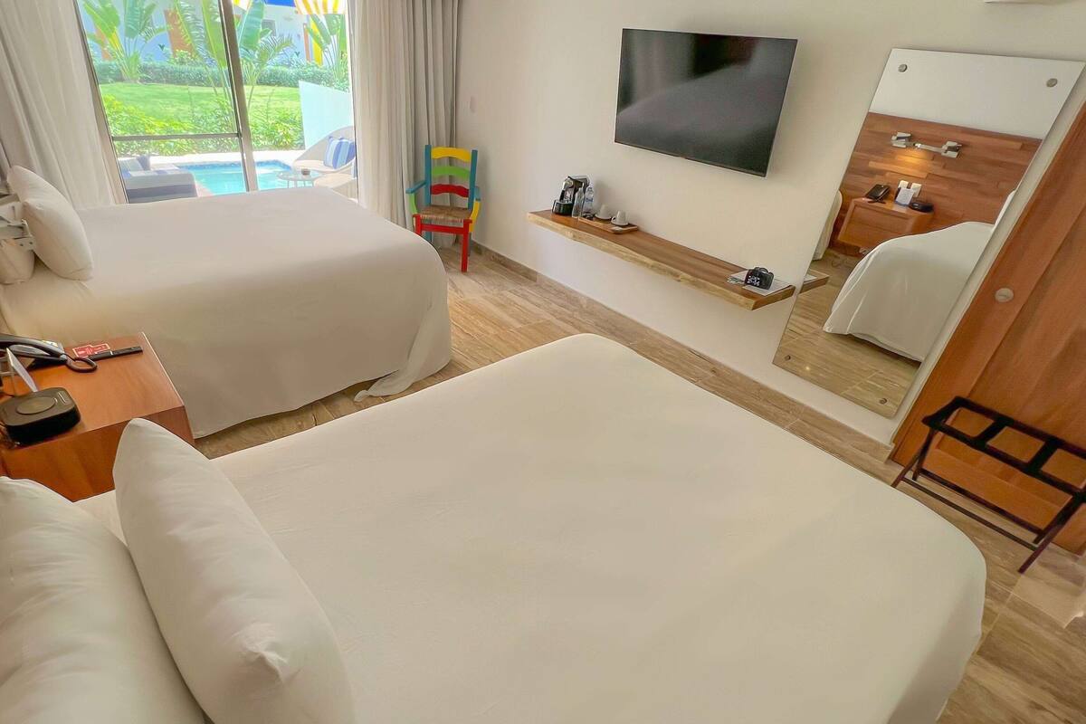 A hotel room with two beds, a TV, a small table with a phone and a remote, and a window with a view of a pool and greenery.