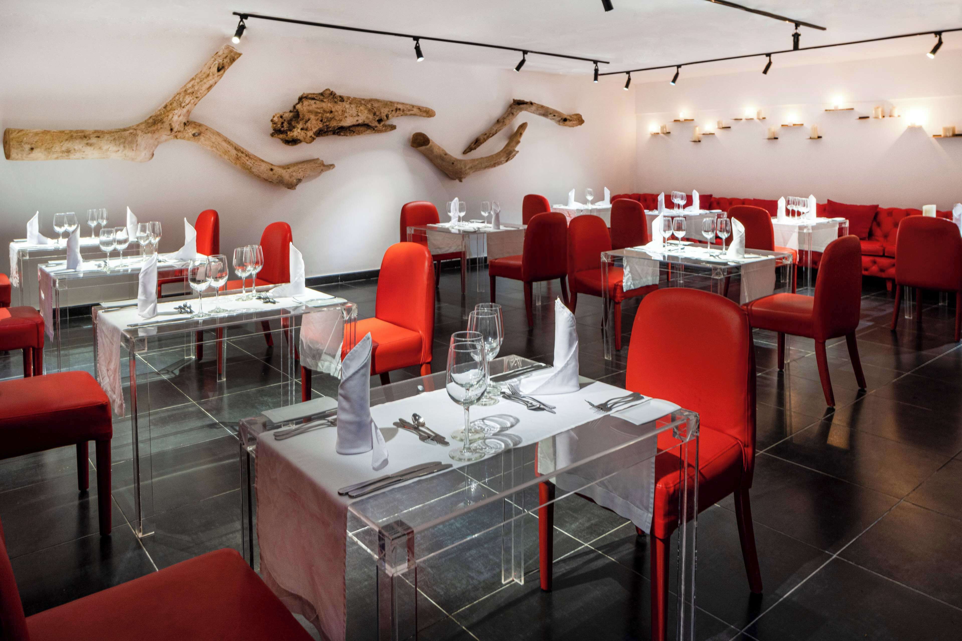 A modern restaurant interior with clear glass tables, red chairs, and a unique wooden wall decor.