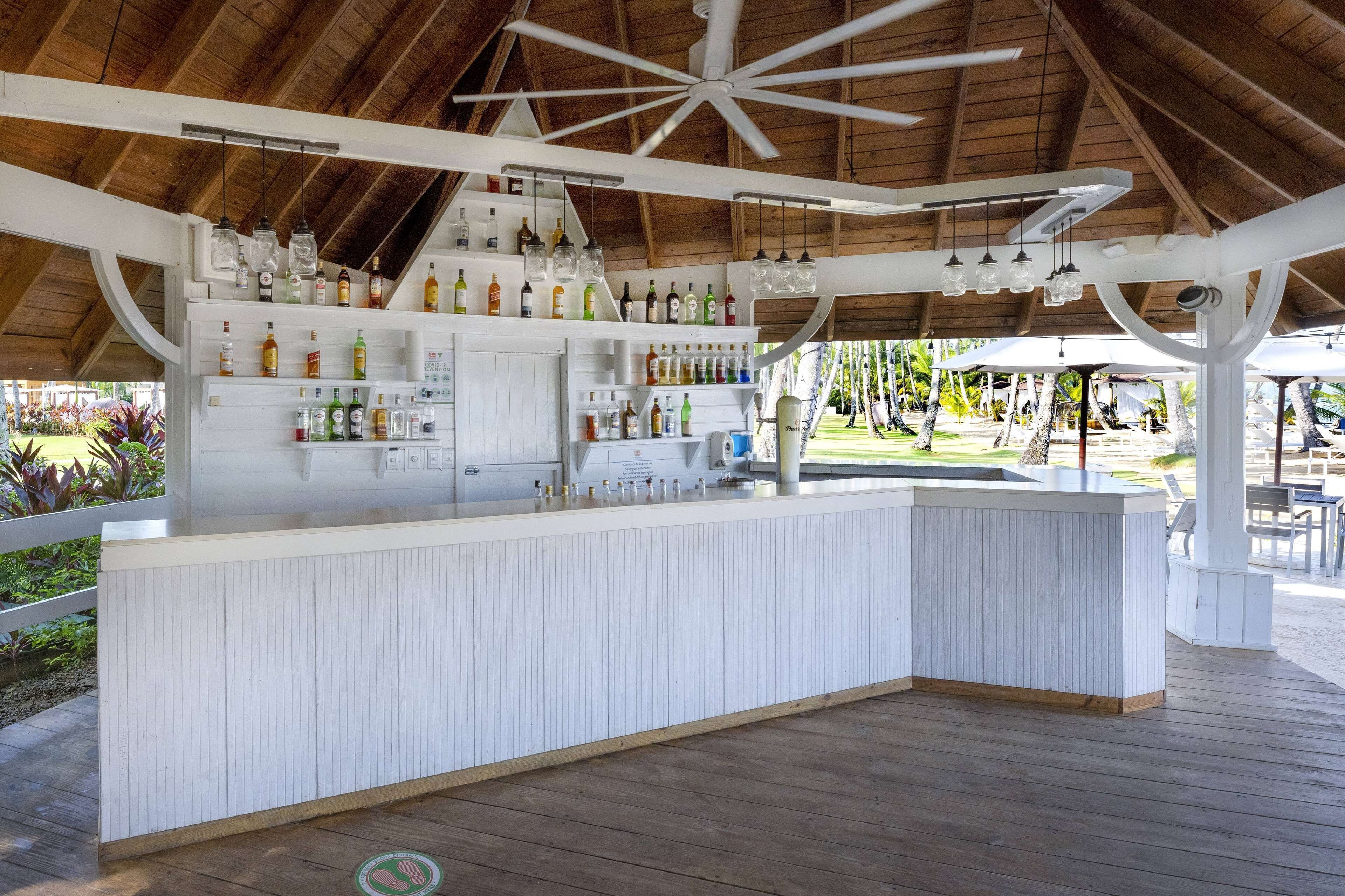 Zona de bar con techo de madera, barra blanca y diferentes botellas y vasos.