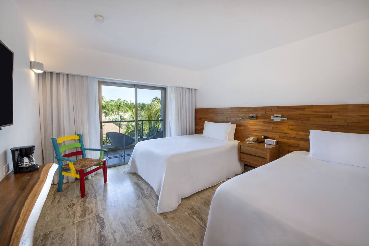 A hotel room with two beds, a wooden headboard, a chair, a coffee maker, and a view of palm trees through the window.