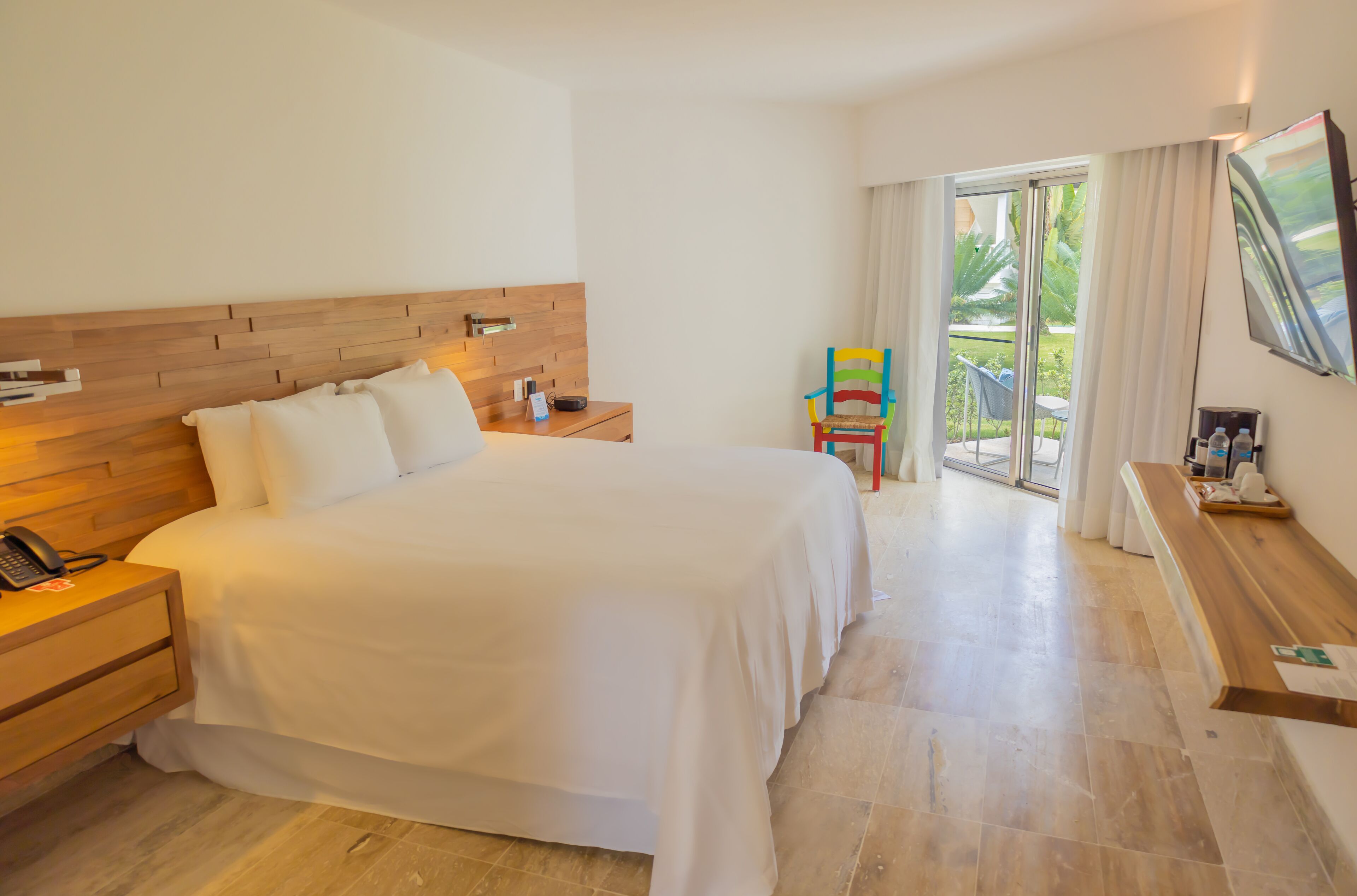 A hotel room with a large bed, wooden headboard, bedside tables, a chair, a TV, and a balcony with curtains.