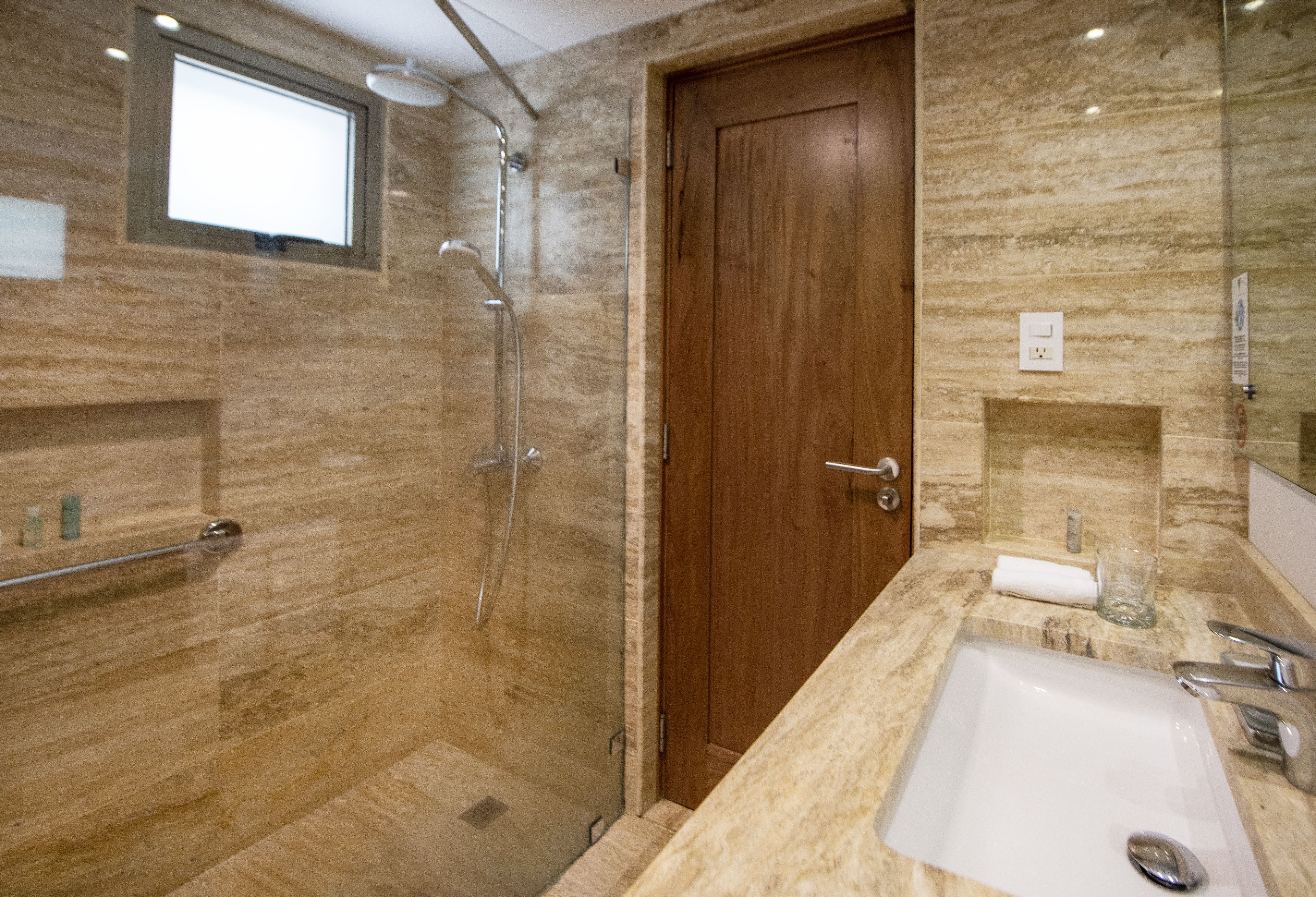 Un baño moderno con cabina de ducha de vidrio, puerta de madera, encimera de mármol y lavabo blanco.