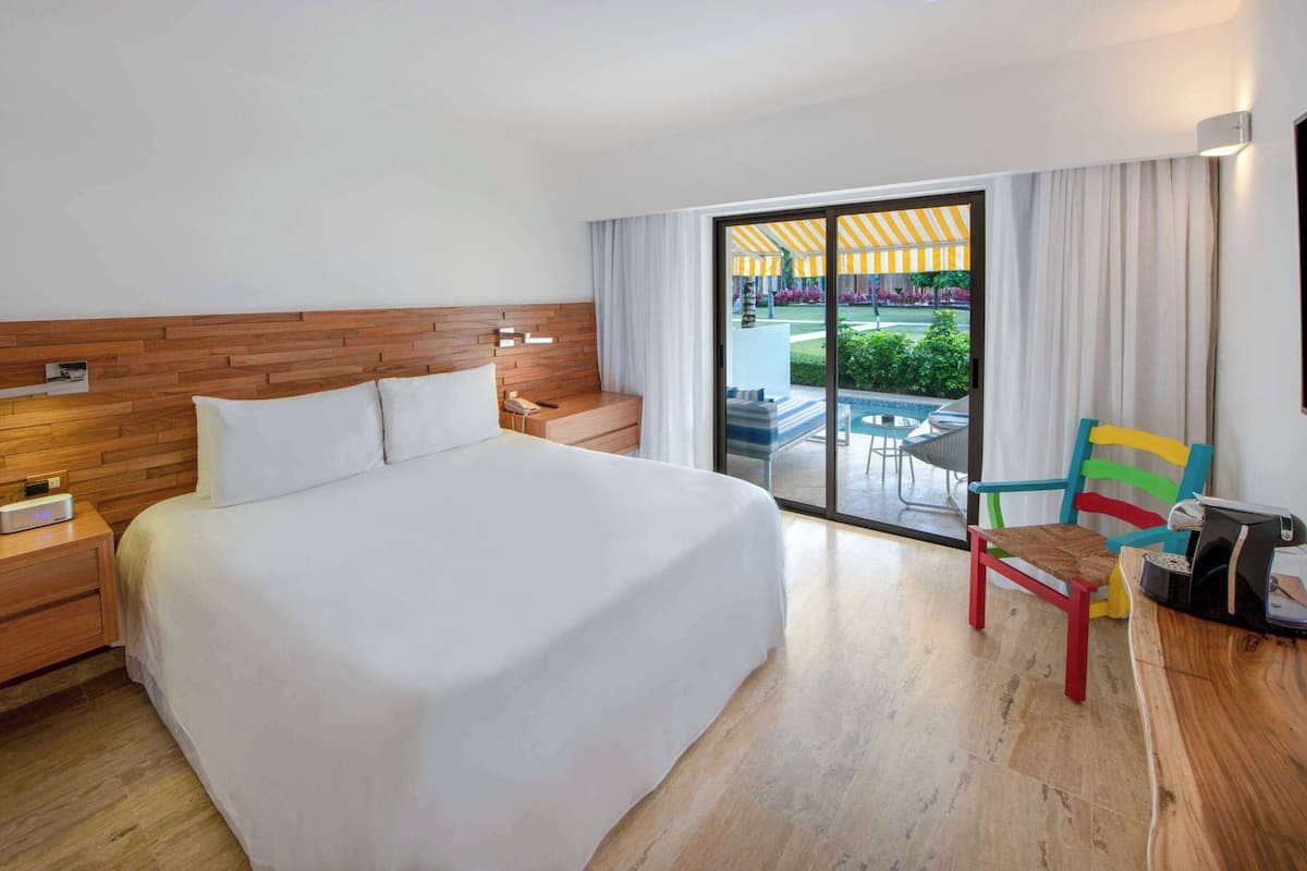 A hotel room with a large bed, a wooden headboard, a nightstand, a desk with a chair, and a view of a pool area through a sliding glass door.