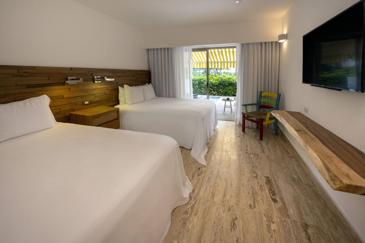 A hotel room with two beds, a wooden headboard, a TV mounted on the wall, and a view of greenery outside.