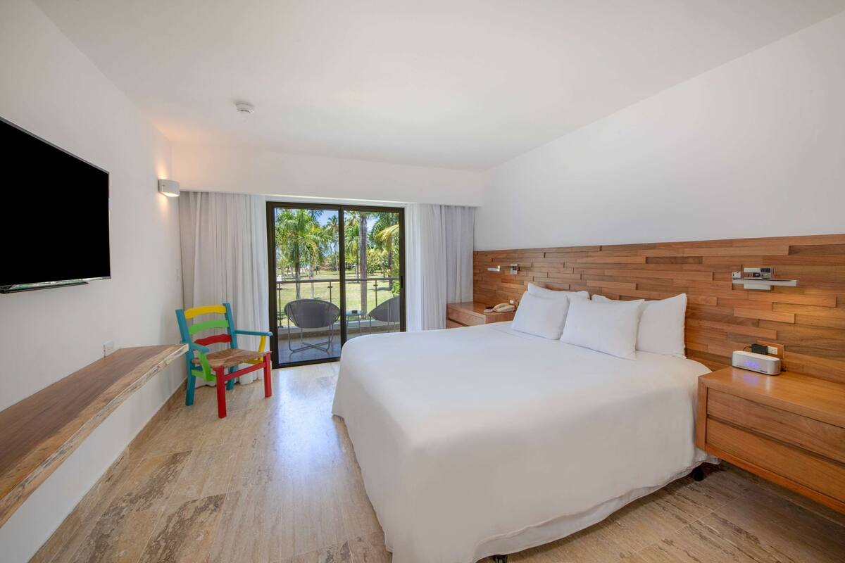 A hotel room with a large bed, a wooden headboard, a flat-screen TV, a colorful chair, and a view of the outdoors through a glass door.