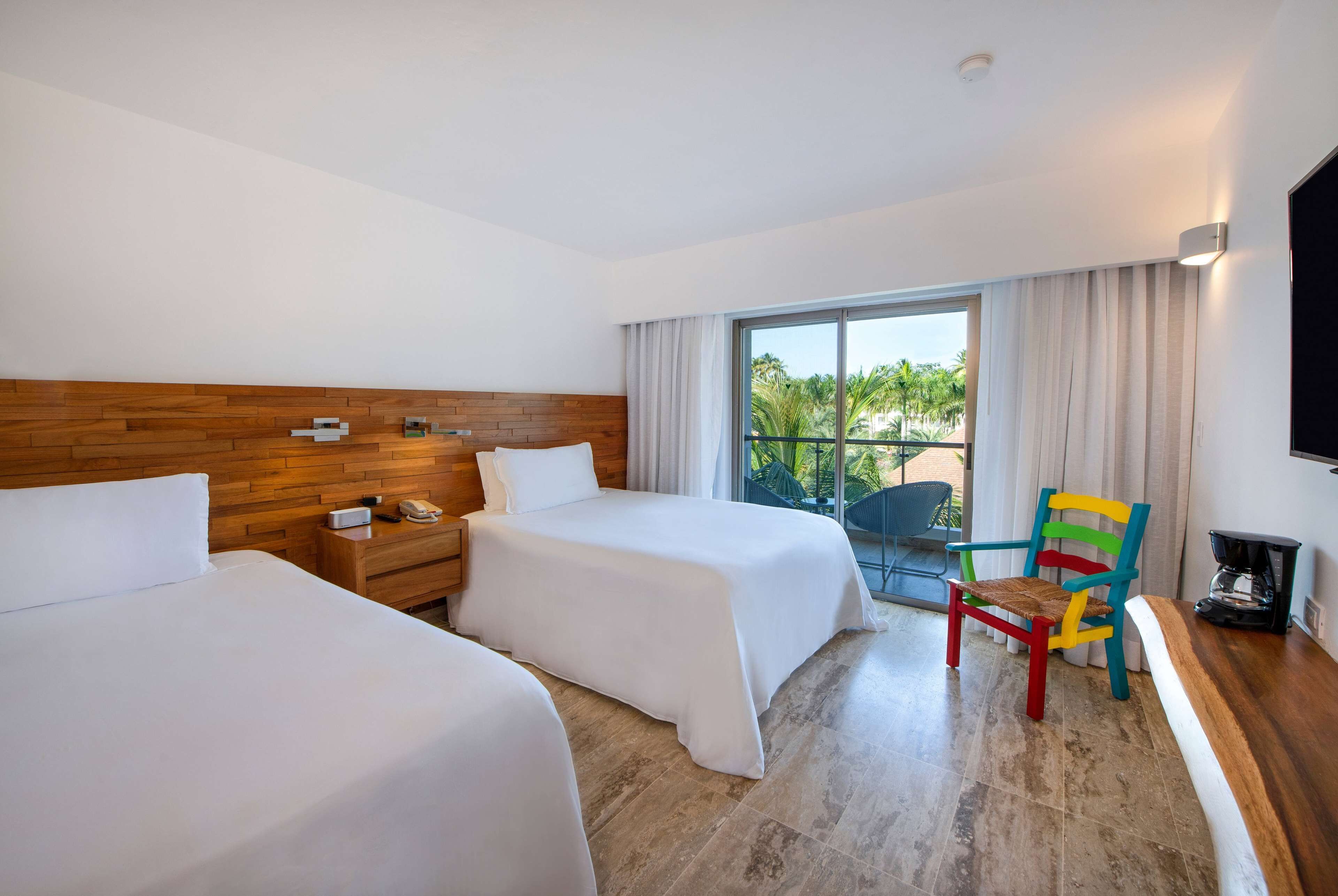 Une chambre d’hôtel avec deux lits, une tête de lit en bois, une petite table avec un téléphone, une chaise colorée et un balcon avec vue sur les palmiers.