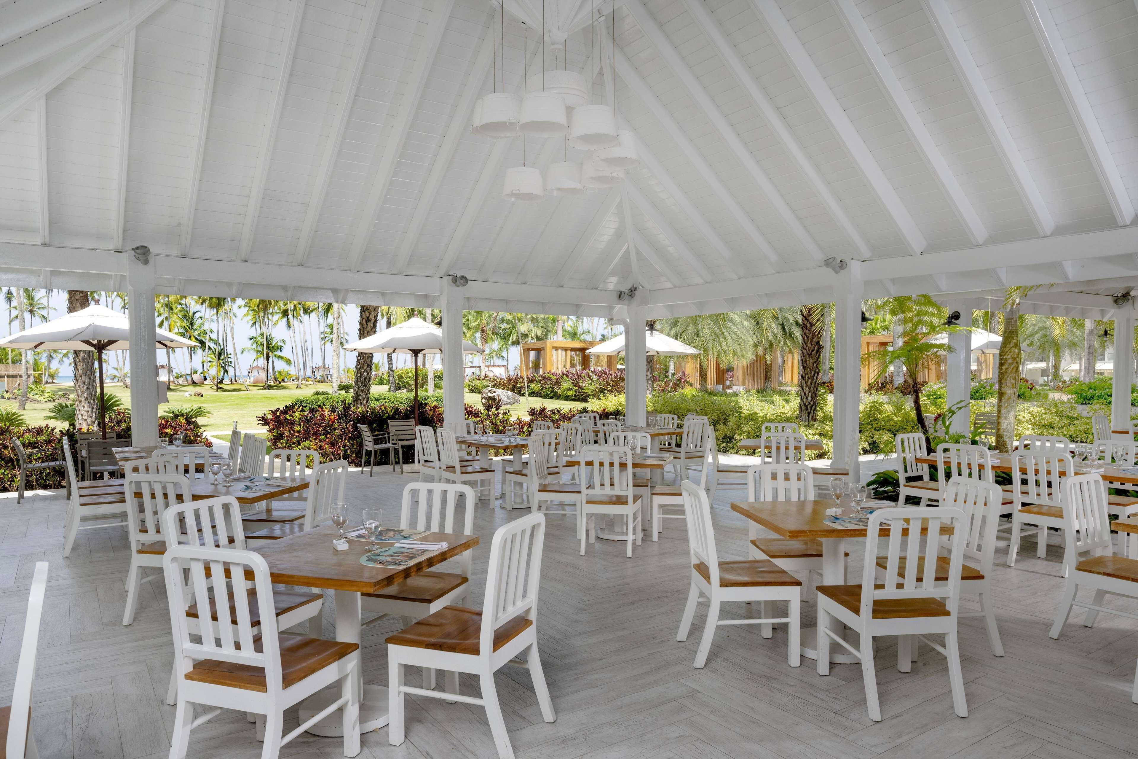 Un espace extérieur abrité, aménagé pour les repas, avec des tables et des chaises blanches, entouré de palmiers et de verdure.