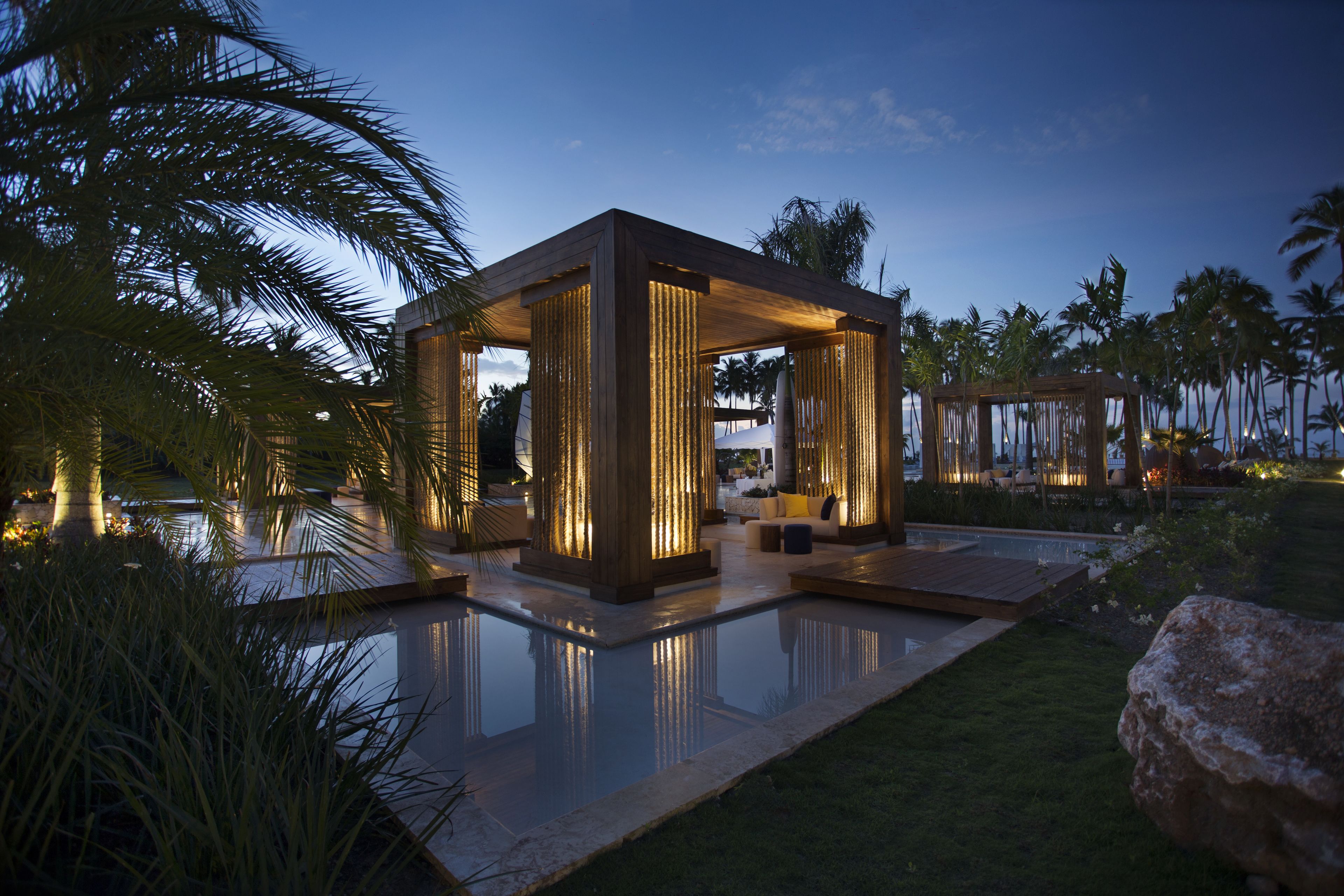 A modern outdoor lounge area with a pool, wooden pergolas, and palm trees.